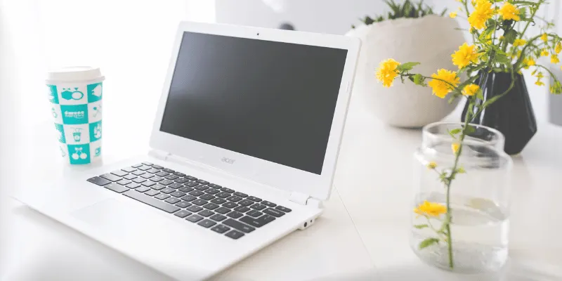 Open white Chromebook beside a coffee cup and yellow flowers on a clean desk
